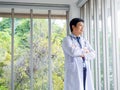 Smiling Asian man doctor portrait in white coat standing alone with crossed arms near glass windows in medical office. Royalty Free Stock Photo