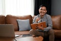 Smiling Asian man making list or taking notes on his notebook while sitting on sofa Royalty Free Stock Photo