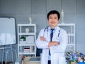 Smiling Asian man orthopedic doctor portrait in white coat standing with crossed arms near bookshelf. Royalty Free Stock Photo