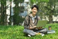 Smiling asian man student using laptop on green grass in front of university building. Education, technology and