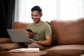 Smiling Asian man using his smartphone and laptop while sitting on a sofa in his living room Royalty Free Stock Photo