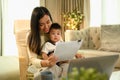 Smiling Asian mother working at home with laptop and documents sitting on armchair with cute little baby son Royalty Free Stock Photo