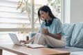 Smiling asian woman in headphones using laptop computer while having her morning coffee sitting on a couch at home Royalty Free Stock Photo