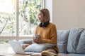 Smiling asian woman in headphones using laptop computer while having her morning coffee sitting on a couch at home Royalty Free Stock Photo