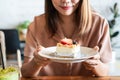 Smiling Asian woman holding a plate of her favorite strawberry cheese cake on wooden table in cafe. Food and dessert concept