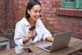 Smiling asian woman with laptop and wireless earphones, paying with credit card, buying online, sitting with cup of Royalty Free Stock Photo