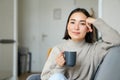 Smiling asian woman sitting on sofa with her mug, drinking coffee at home and relaxing after work, looking calm and cozy Royalty Free Stock Photo