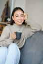 Smiling asian woman sitting on sofa with her mug, drinking coffee at home and relaxing after work, looking calm and cozy Royalty Free Stock Photo