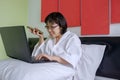 Smiling Asian woman in white bathrobe talking on smartphone and using laptop on her bed at morning time Royalty Free Stock Photo