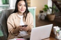 Smiling asian woman working with laptop from home. Girl student holding smartphone, sitting near computer and looking at Royalty Free Stock Photo