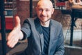 Smiling attractive adult successful bald bearded man in suit with laptop giving handshake, hand of help, greeting at cafe Royalty Free Stock Photo