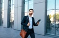 Smiling attractive european millennial bearded businessman in suit walks with bag and tablet on street Royalty Free Stock Photo