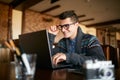Smiling Attractive Hipster Man In Glasses And Winter Sweater Working On Laptop In Cafe. Freelancer Businessman Browsing