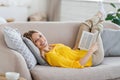 Smiling attractive millennial blond european lady in yellow clothes lies on sofa and reads book in living room
