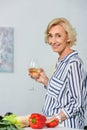 smiling attractive senior woman holding glass of white wine in kitchen and looking Royalty Free Stock Photo