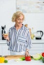 smiling attractive senior woman holding glass of white wine in kitchen and looking Royalty Free Stock Photo