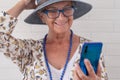 Smiling attractive senior woman wearing blue hat using mobile phone - standing against a white brick wall - elderly people Royalty Free Stock Photo