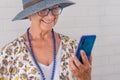 Smiling attractive senior woman wearing blue hat using mobile phone - standing against a white brick wall - elderly people Royalty Free Stock Photo