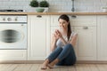 Smiling attractive young woman using smartphone sitting on kitchen floor. Royalty Free Stock Photo