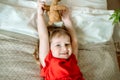 Smiling baby lies on a bed with a teddy bear in a red dress..Cute red-haired girl plays at home in a bright bedroom. funny and