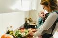 Smiling baby ported in baby carrier backpack looking at his mother while she cooks, concept of family conciliation