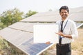 Smiling bank officer showing white empty board by looking at camera in front of solar panal at farmland - concept of