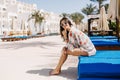 Smiling barefooted girl in stylish clothes sitting on blue chaise-longue and enjoying tropical sun. Adorable laughing