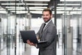 Smiling bearded business man focused on company datas while standing in office corridor and using laptop. Royalty Free Stock Photo