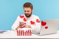 Smiling bearded businessman in white shirt in red heart shaped stickers sitting at desk with laptop and striped giftbox typing on Royalty Free Stock Photo