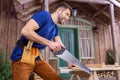 Smiling bearded carpenter in tool belt sawing wooden plank on porch