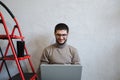 Smiling bearded hipster using laptop on white background near red mettalic stairs.