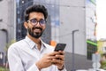 Smiling bearded young man in glasses using smartphone in urban setting, exuding confidence and connectivity Royalty Free Stock Photo