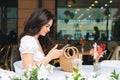 Smiling beautiful brunette young woman in white clothes using mobile phone with straw bag on table at terrace of the summer cafe Royalty Free Stock Photo