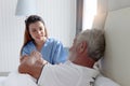 Smiling beautiful caring female doctor holding hand of male senior patient who lying in hospital bed. Nurse takes care elderly man