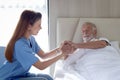 Smiling beautiful caring female doctor holding hand of male senior patient who lying in hospital bed. Nurse takes care elderly man Royalty Free Stock Photo