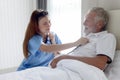 Smiling beautiful caring female doctor using stethoscope listen to senior patient heart who lying in hospital bed. Nurse takes