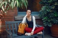 Smiling beautiful Caucasian young girl woman in white sweater and red jeans, sitting with yellow travel bag backpack on bench