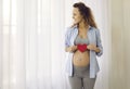 Smiling beautiful future young mother holding red heart near big pregnant belly. Royalty Free Stock Photo