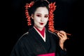 Beautiful geisha in black kimono with red flowers in hair holding chopsticks isolated on black Royalty Free Stock Photo