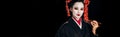 Beautiful geisha in black kimono with red flowers in hair holding chopsticks isolated on black, panoramic shot Royalty Free Stock Photo