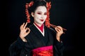 Beautiful geisha in black kimono with red flowers in hair holding chopsticks and showing okay sign isolated on black Royalty Free Stock Photo