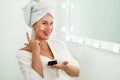 Smiling beautiful lady apply skincare cream on face look in bathroom mirror, happy young woman wrap towel on head put Royalty Free Stock Photo