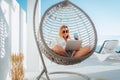 Smiling beautiful middle-aged woman sitting in a comfortable hanging chair on the open house terrace with a glass of water and Royalty Free Stock Photo