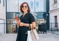 Smiling beautiful modern middle-aged female Portrait dressed black dress and sunglasses with shopping bags holding slim smartphone