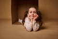 Smiling beautiful positive pleasant baby girlin pajamas with two ponytails inside a cardboard box, looking at camera isolated over