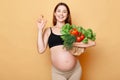 Smiling beautiful pregnant woman holding fresh vegetables posing isolated over beige background crossing fingers hopes buying Royalty Free Stock Photo