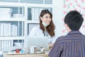 Smiling beautiful young Asian female doctor with white coat is using laptop computer and talking to handsome male patient about Royalty Free Stock Photo