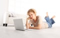 smiling beautiful young woman with computer laptop, lying on living room wooden floor in modern comfortable white home Royalty Free Stock Photo