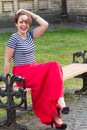 Smiling beautiful young woman with short hear in vest and long red skirt, sitting on a bench outdoor Royalty Free Stock Photo