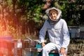 Smiling beekeeper in white uniform. Man sitting near work equipment and beehives.